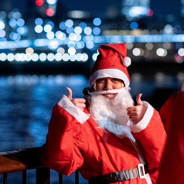 Person dressed as Santa Claus giving two thumbs up, smiling in front of a night-time cityscape with festive lights reflecting on the water.
