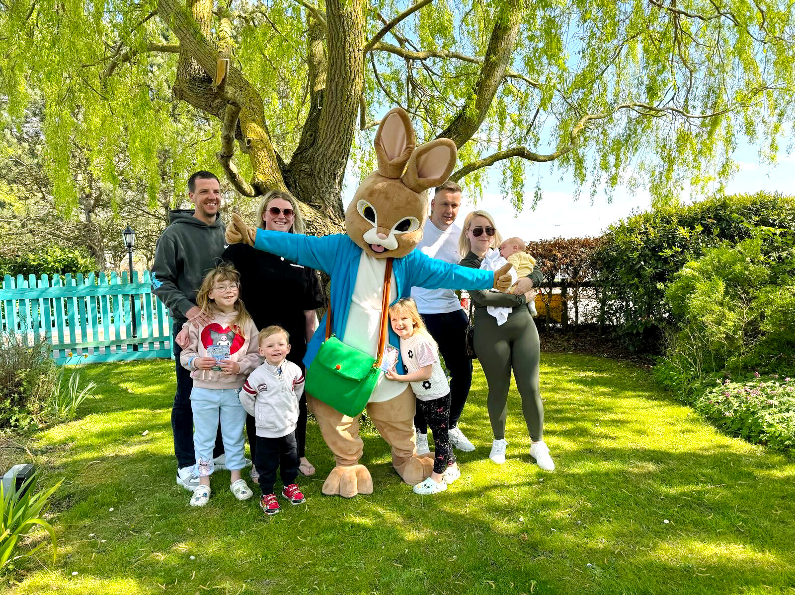Group of adults and children posing outdoors with a person in a bunny costume under a large tree in a sunny park setting