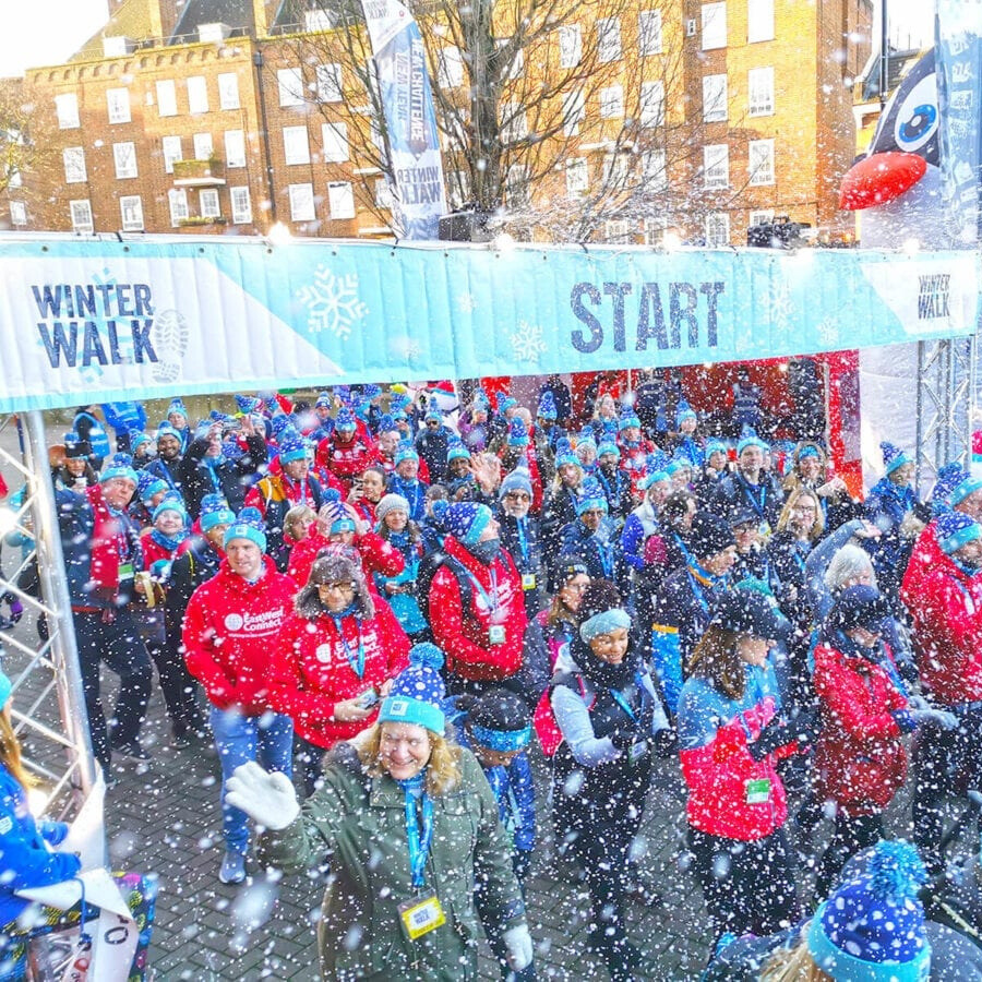 Participants gathered at the starting line of the Winter Walk event, dressed in winter clothing with artificial snow falling, set in an urban outdoor location with festive atmosphere.