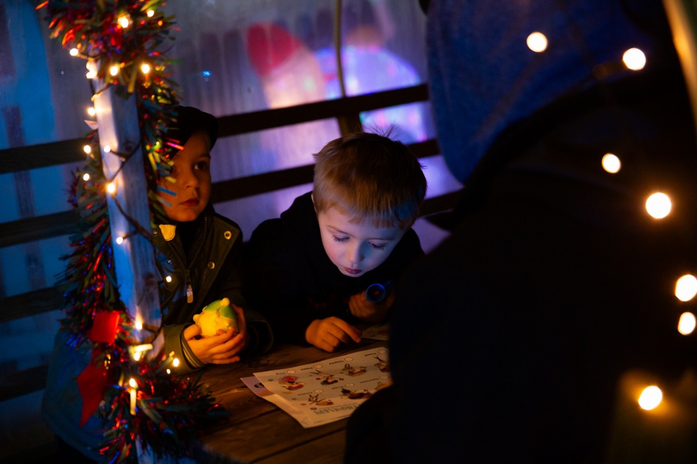Two children sitting at a festively decorated table with string lights, one holding a yellow object and the other focused on a piece of paper, in a cozy, holiday-themed setting.