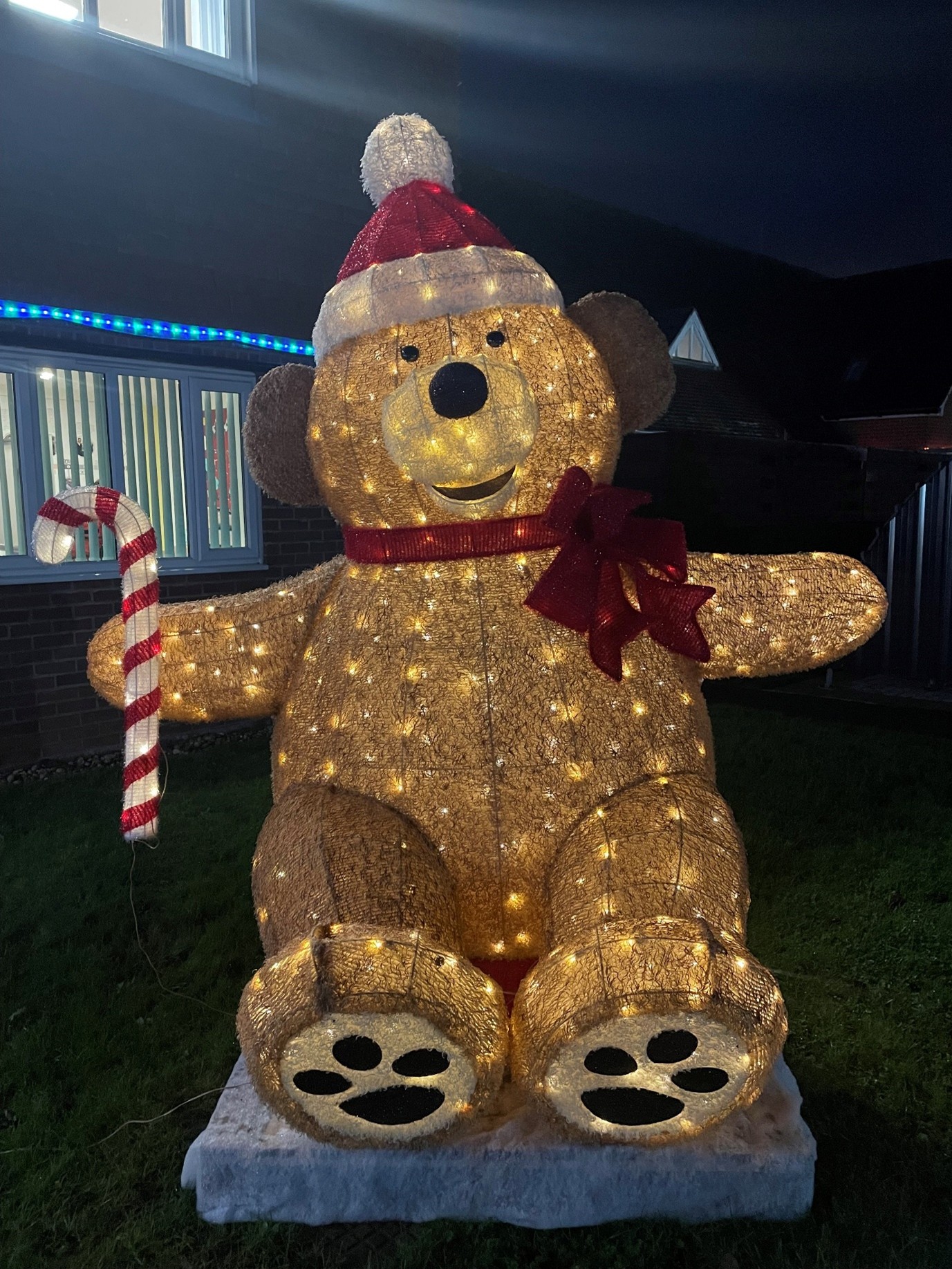 Large illuminated teddy bear Christmas decoration wearing a Santa hat and red ribbon, holding a candy cane, displayed outdoors at night with festive blue lights and window blinds in the background