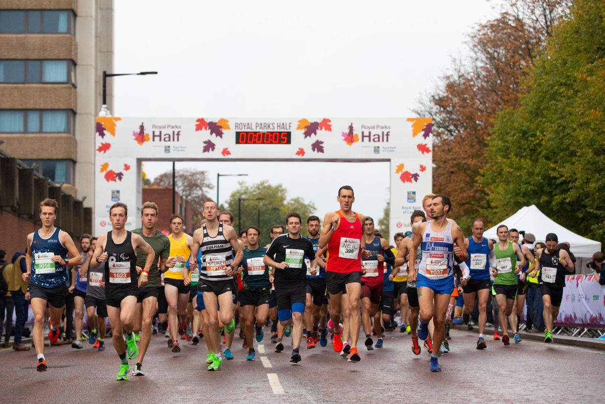 Runners at the start line of the Royal Parks Half marathon, with a banner displaying autumn leaves and a digital timer reading 00:00:05, set against a backdrop of trees with fall foliage and nearby buildings.