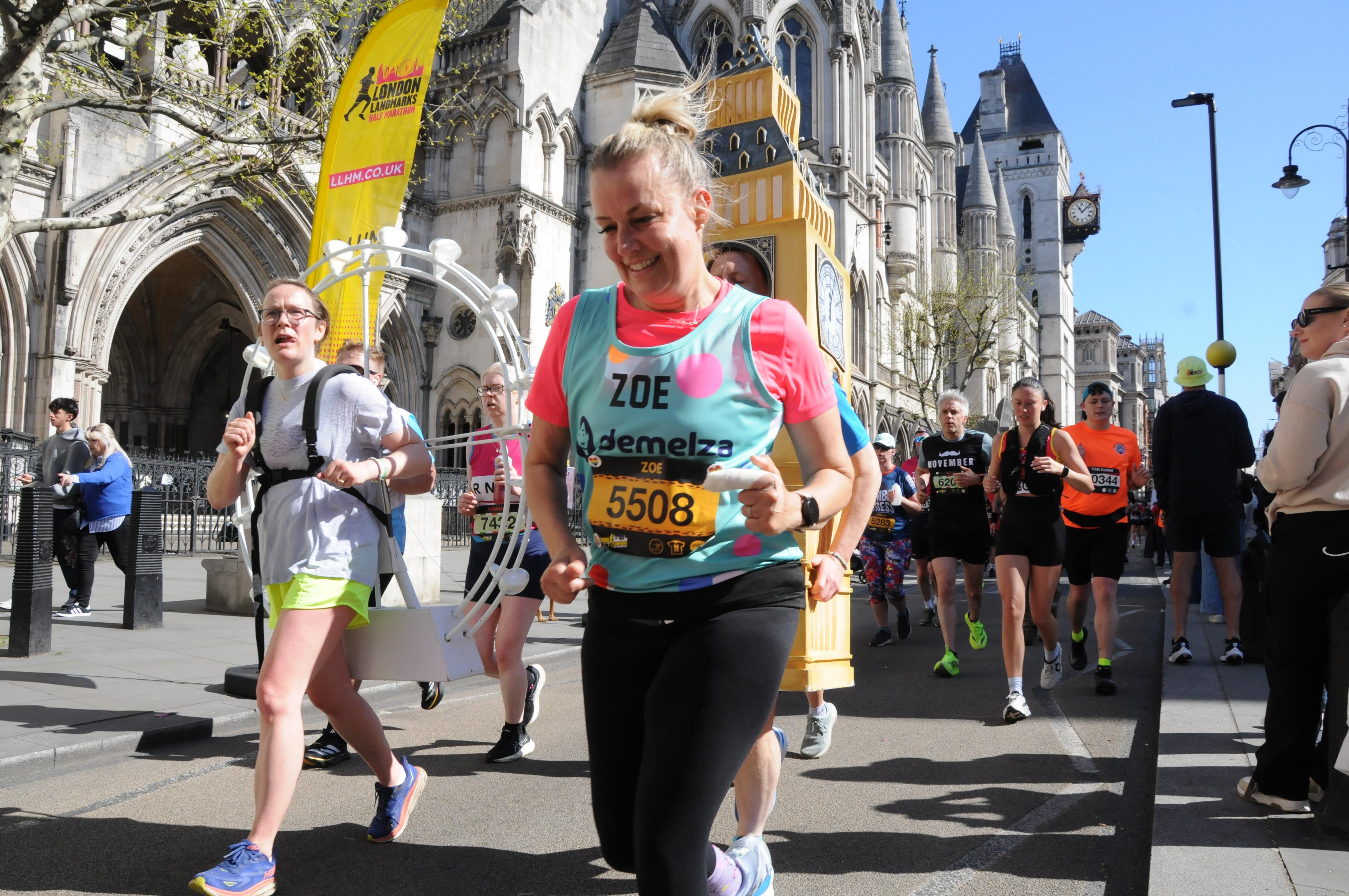 Participants running in the London Marathon, with one wearing a turquoise 'Zoe – Demelza' vest and bib number 5508, and another dressed as Big Ben, set against historic buildings and a yellow event banner.