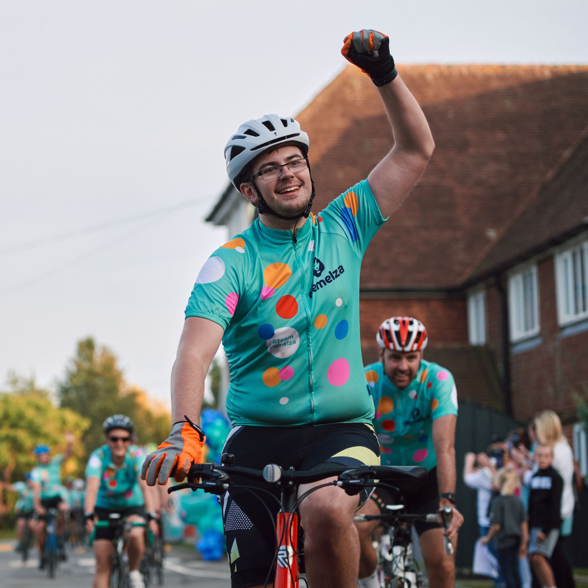 Cyclist wearing a polka-dot 'Demelza' jersey raising one arm in celebration during a group cycling event, with fellow riders and spectators lining a residential street.