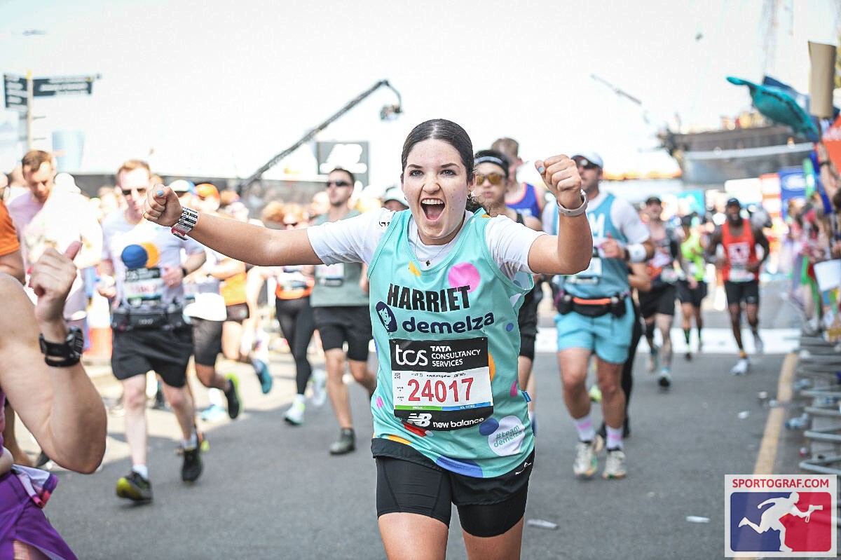 Runner named Harriet wearing a 'Demelza' shirt with bib number 24017, arms raised in celebration during a marathon event, surrounded by fellow participants and branded with TCS and New Balance logos.