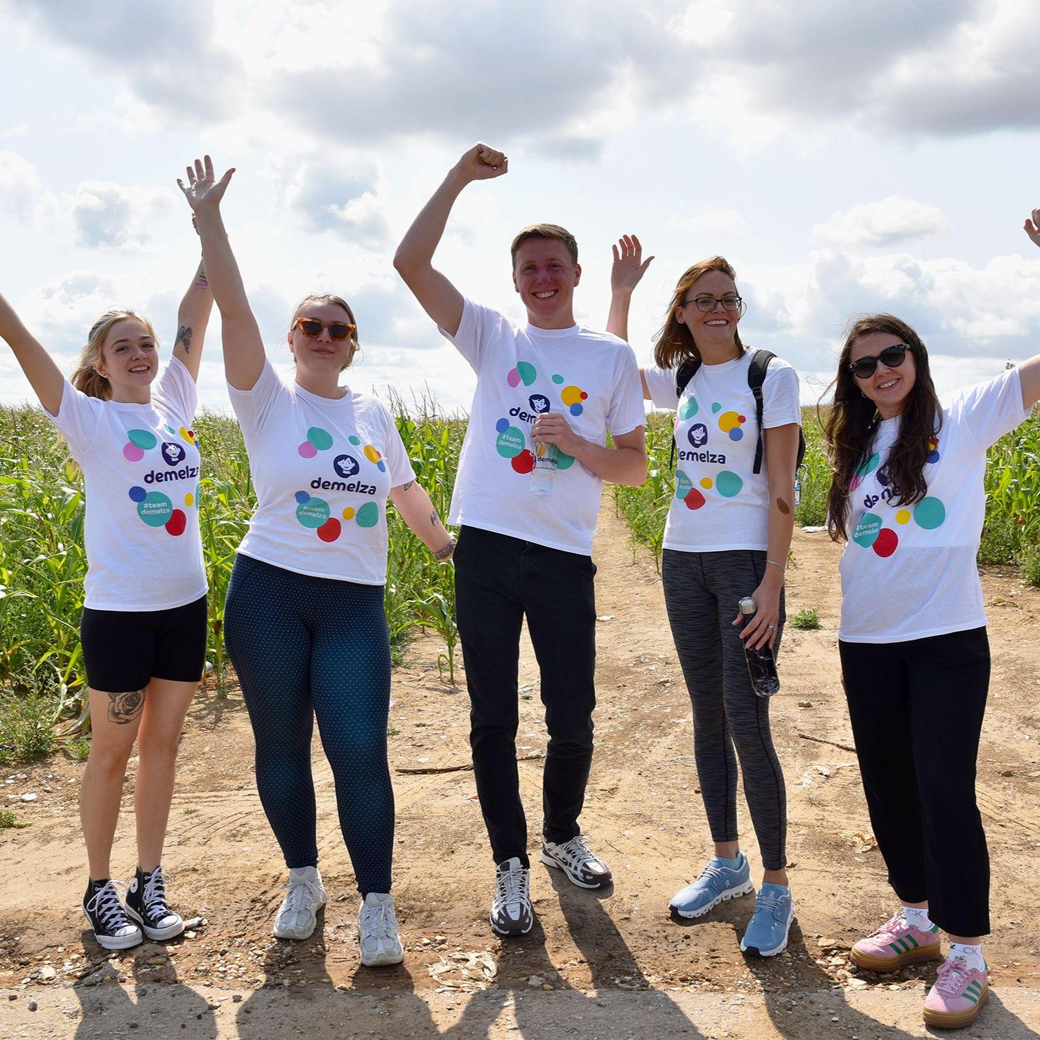Group of five people celebrating outdoors in Demelza charity t-shirts, standing on a dirt path surrounded by greenery under a cloudy sky.