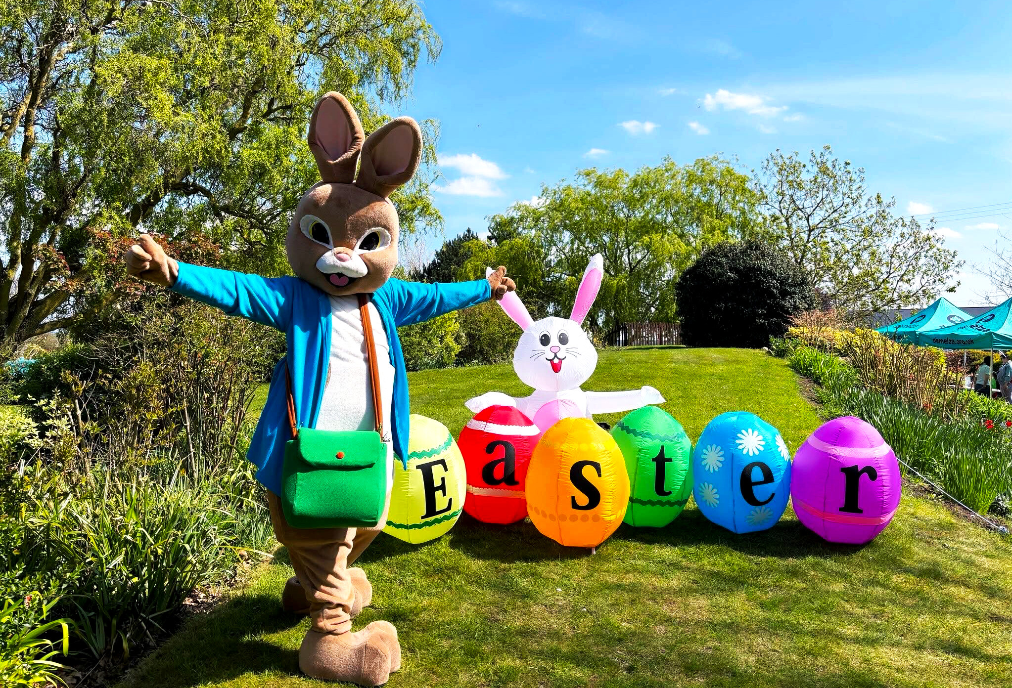 Person in a bunny costume with large ears, blue jacket, and green bag standing beside colorful Easter eggs spelling 'Easter', with an inflatable white bunny in a grassy outdoor setting under a bright blue sky.