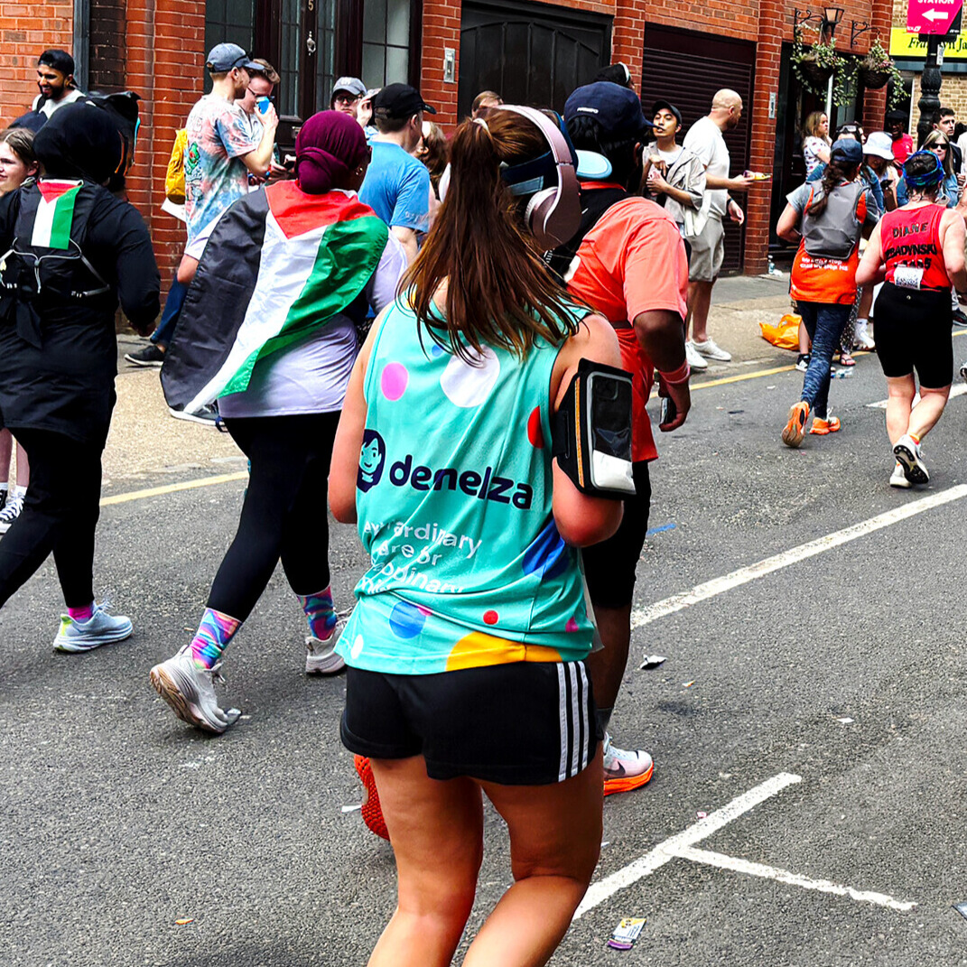 Group of runners participating in a public marathon event, with one wearing a 'Demelza – Ordinary to Extraordinary' shirt, flags draped over shoulders, and spectators lining a street of brick buildings.