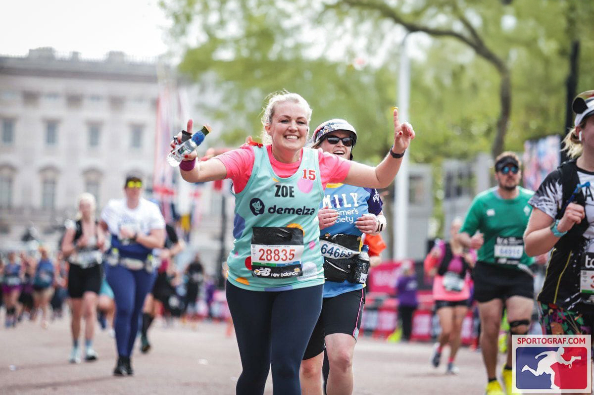 Runner named Zoe wearing a light blue and pink outfit with bib number 28835, celebrating mid-race during a marathon in an urban setting with fellow participants and trees in the background.