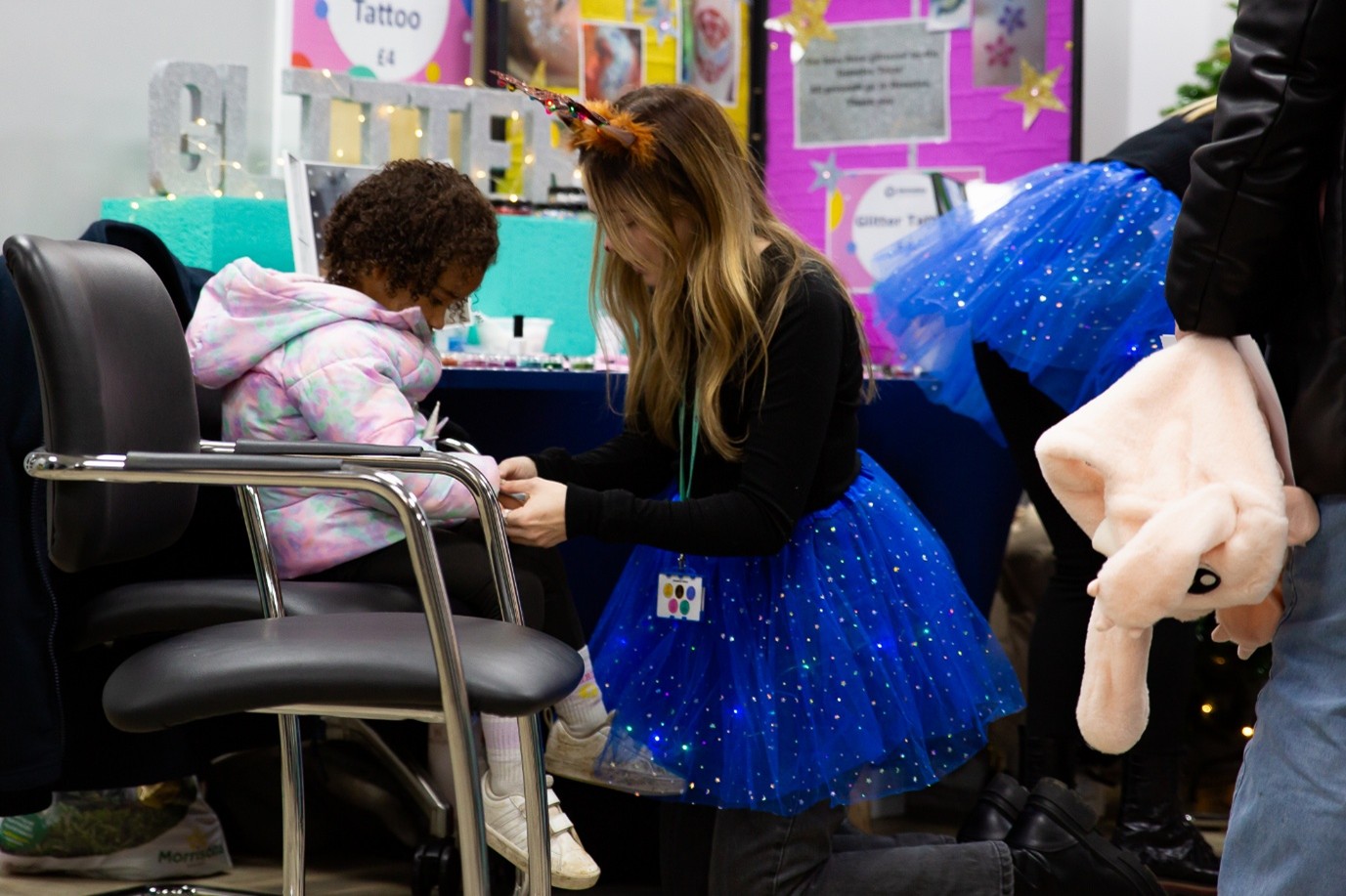 Person in a blue sparkly tutu and antler headband kneeling beside a child in a colorful hoodie at a festive event, with glitter decorations, informational posters, and others in matching outfits in the background.