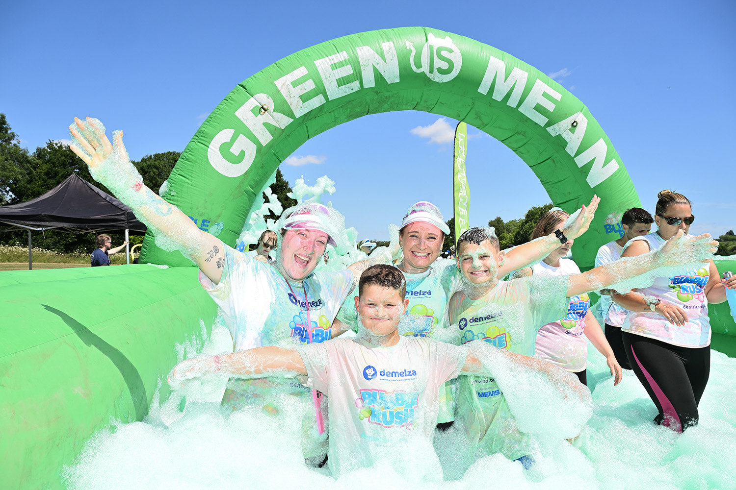 Group of smiling participants covered in foam posing under a 'GREEN IS MEAN' inflatable arch during a sunny outdoor charity event, wearing white t-shirts with colorful logos.
