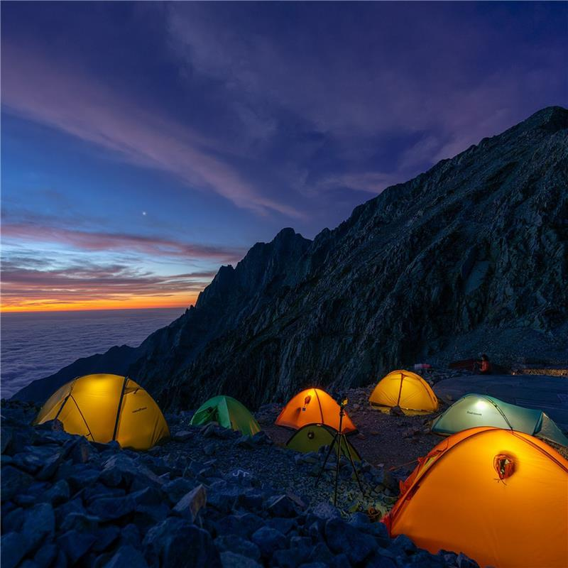 Colorful tents illuminated from within at a twilight mountain campsite, set on a rocky slope with a dramatic sunset sky transitioning from deep blue to warm orange hues.