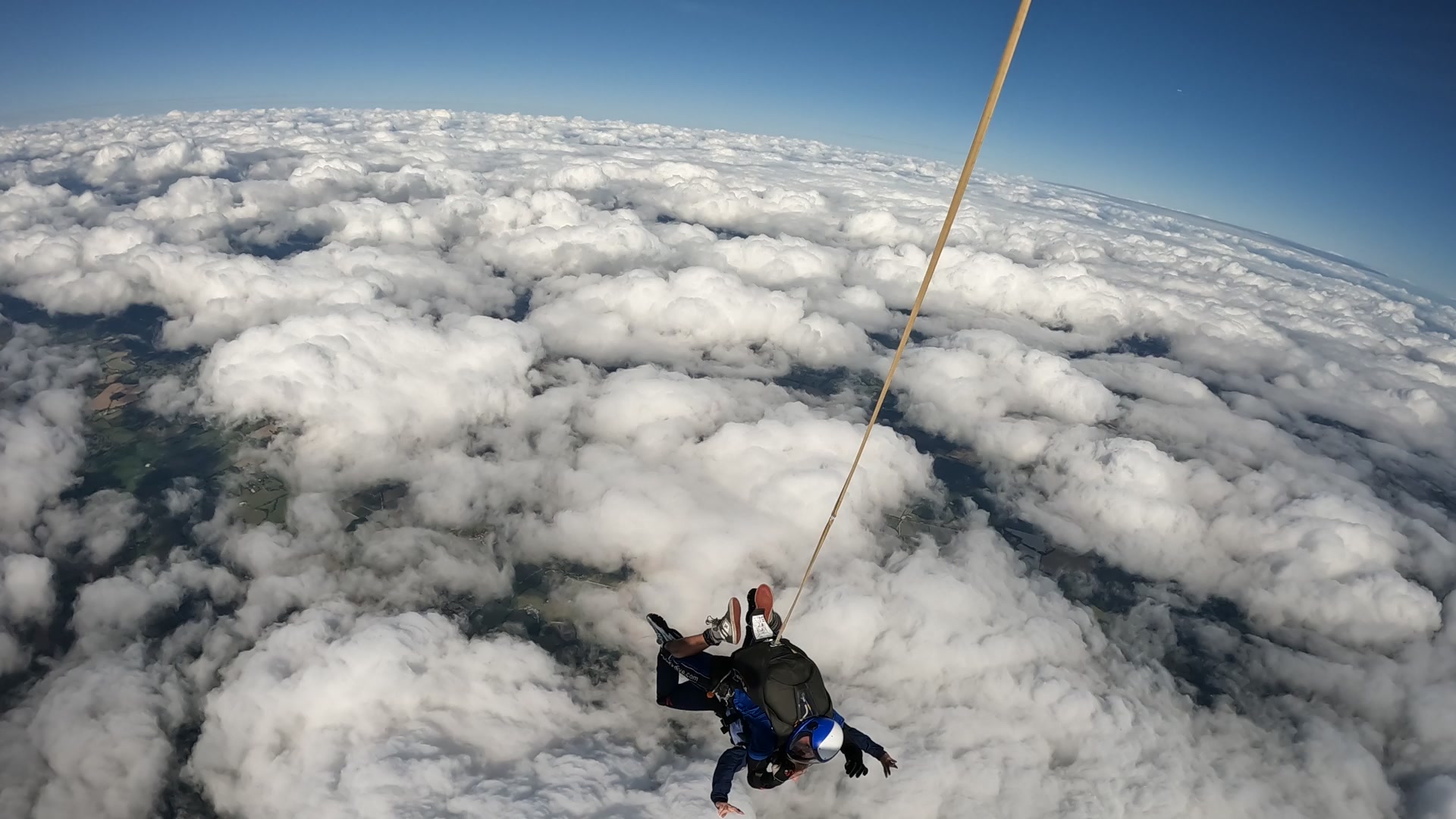 Tandem skydivers in freefall above a dense cloud layer, harnessed together with a yellow cord, set against a clear blue sky.