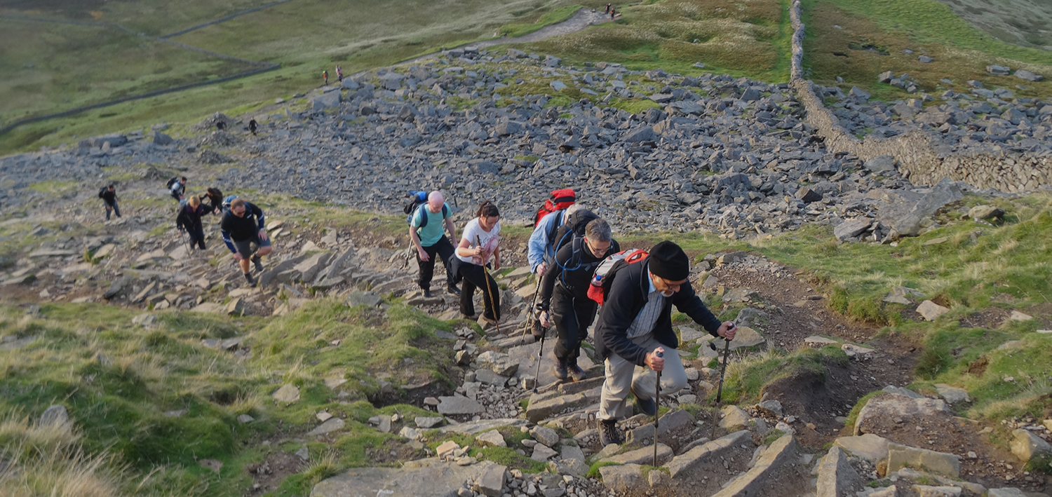 Group of hikers ascending a rocky mountain trail on a steep slope in a rugged landscape, with grassy patches and stone steps leading upward under an open sky.