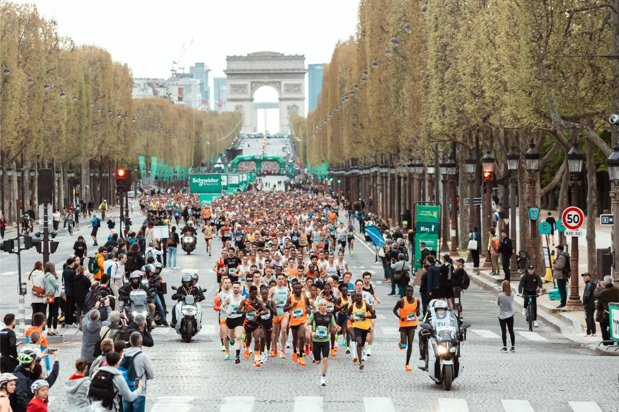 Large group of runners participating in a marathon on a tree-lined avenue in Paris, with the Arc de Triomphe in the background and green event banners lining the street.