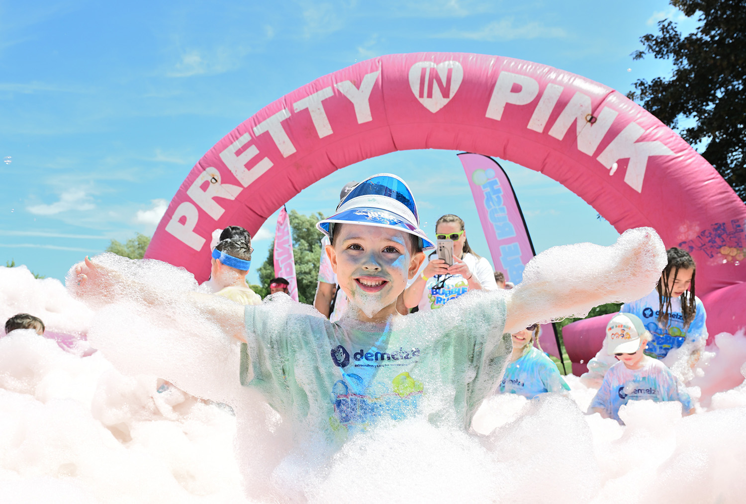 Smiling child wearing a blue visor covered in foam at a sunny outdoor event, standing in front of a large pink inflatable arch labeled 'PRETTY IN PINK', surrounded by other foam-covered participants.