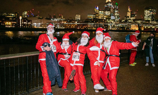 Group of participants in Santa Claus costumes posing at night during a festive fun run, with race numbers and a city skyline illuminated in the background