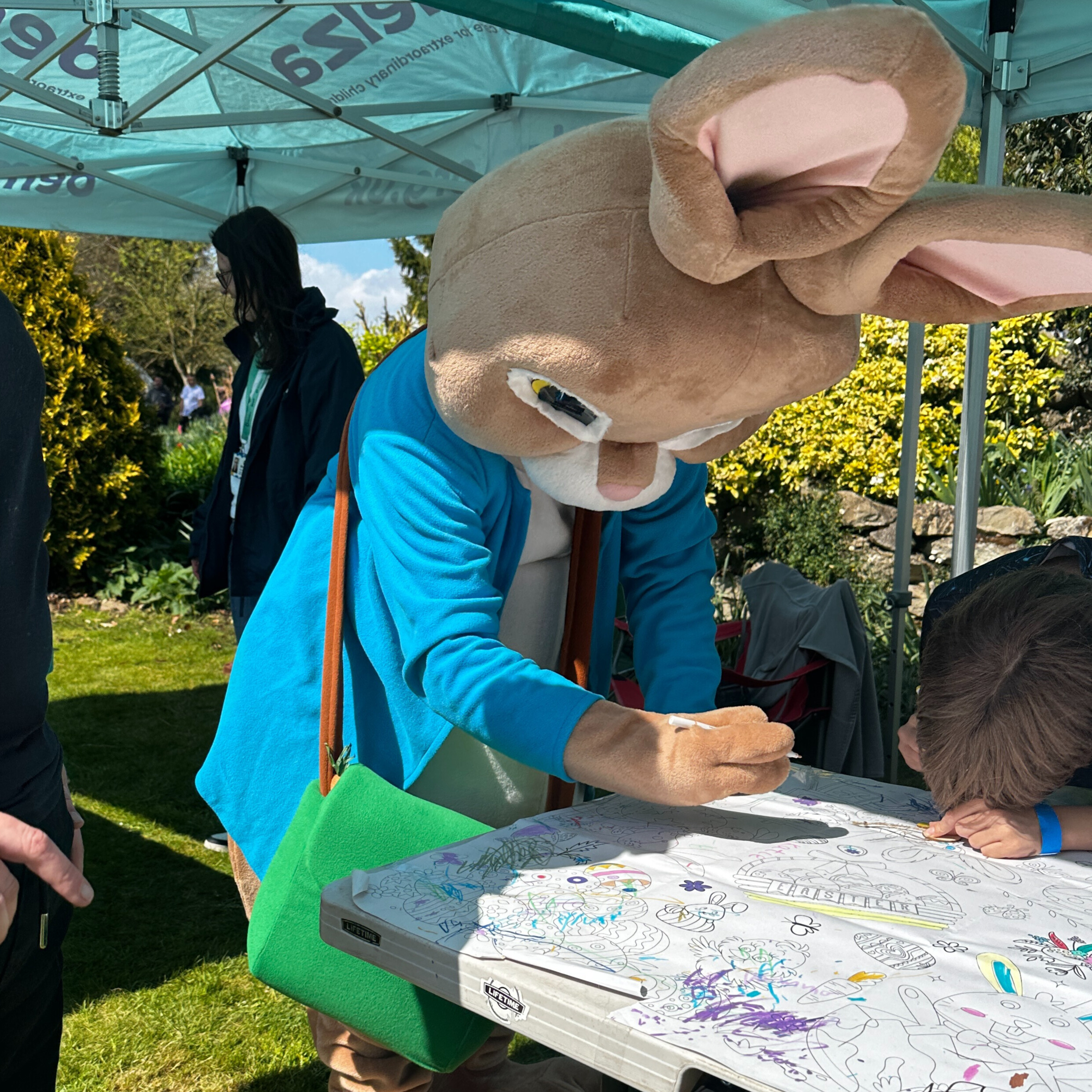 Person in a large rabbit costume with blue jacket and green bag drawing at a table with a child during a family-friendly outdoor event under a tent