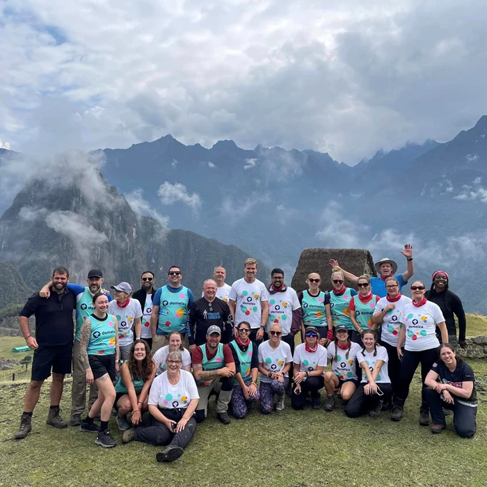 Group of hikers wearing matching white t-shirts posing in front of lush green mountains during a trekking expedition, with cloudy skies and scenic landscape in the background.